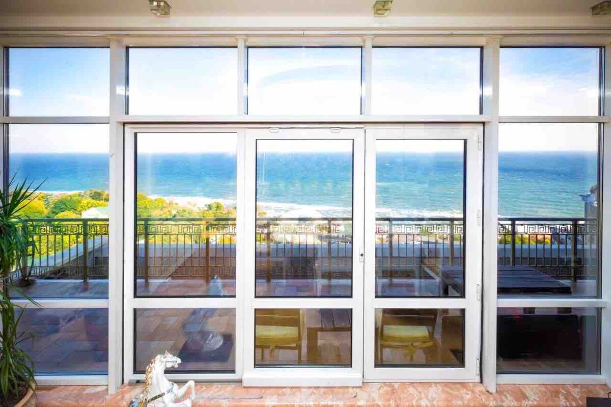 Large glass doors provide an unobstructed view of the sea beyond, framed by a sleek balcony railing. The interior area is brightened by natural light, creating a seamless transition from indoor to outdoor spaces.