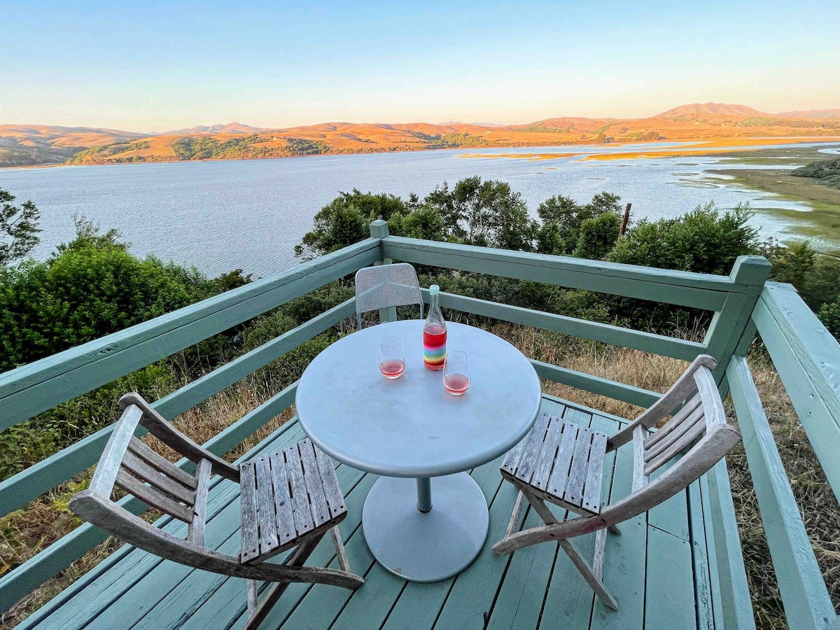 A circular table with two wooden chairs is positioned on a private deck, offering expansive views of Tomales Bay and surrounding hills. The scene captures a tranquil sunset glow, creating a serene atmosphere for outdoor enjoyment.
