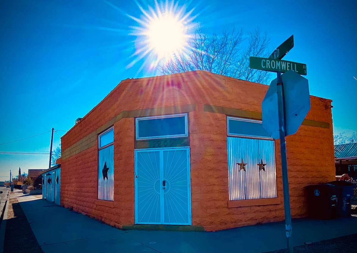 The vibrant exterior of a corner building is depicted, featuring an orange facade adorned with star motifs on the windows. A bright sun shines overhead, creating a lens flare effect. A street sign for Cromwell and another road is visible at the forefront.