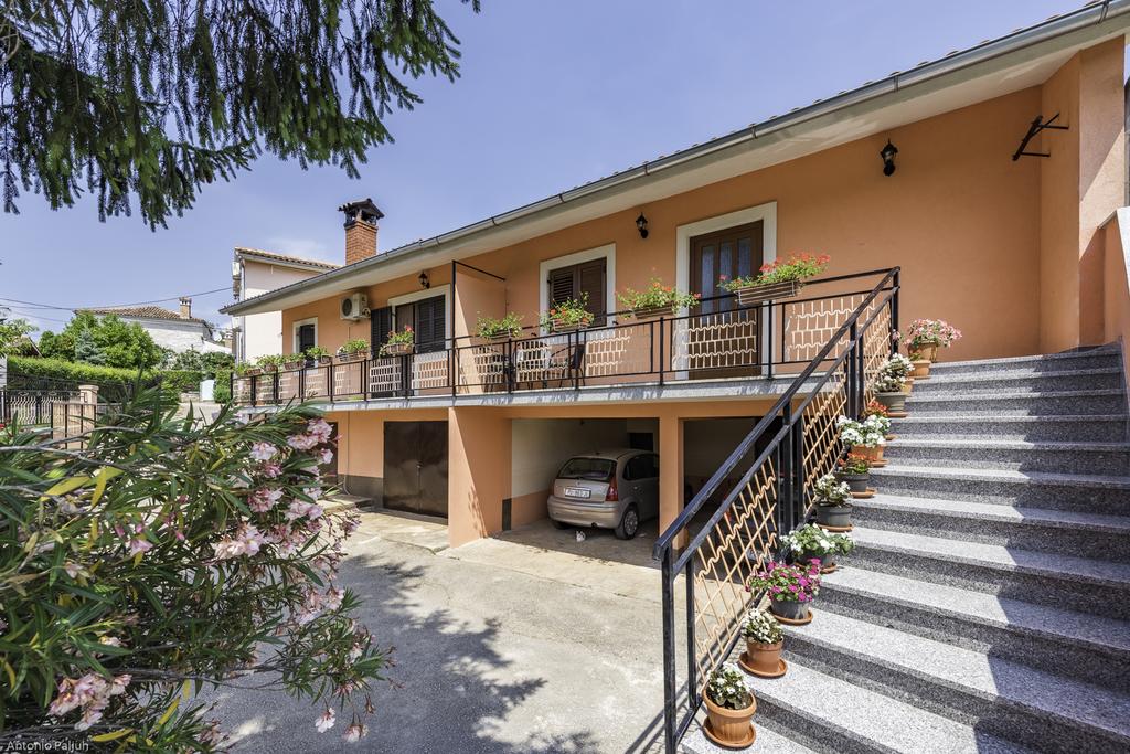 A two-story building features a welcoming façade with a spacious balcony adorned with potted flowers. The entrance is approached by a set of stairs, while a covered garage space is visible at the bottom of the steps. Lush greenery surrounds the property.