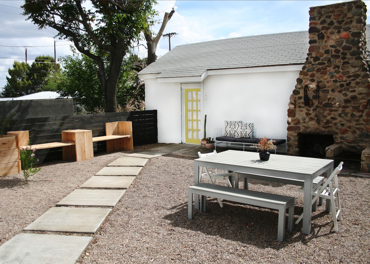 An outdoor courtyard features a mix of gravel and stone pathways leading to seating arrangements. A large picnic-style table is surrounded by white chairs, while a black sofa is positioned near a rustic stone fireplace. A bright yellow door adds a cheerful touch to the white casita.