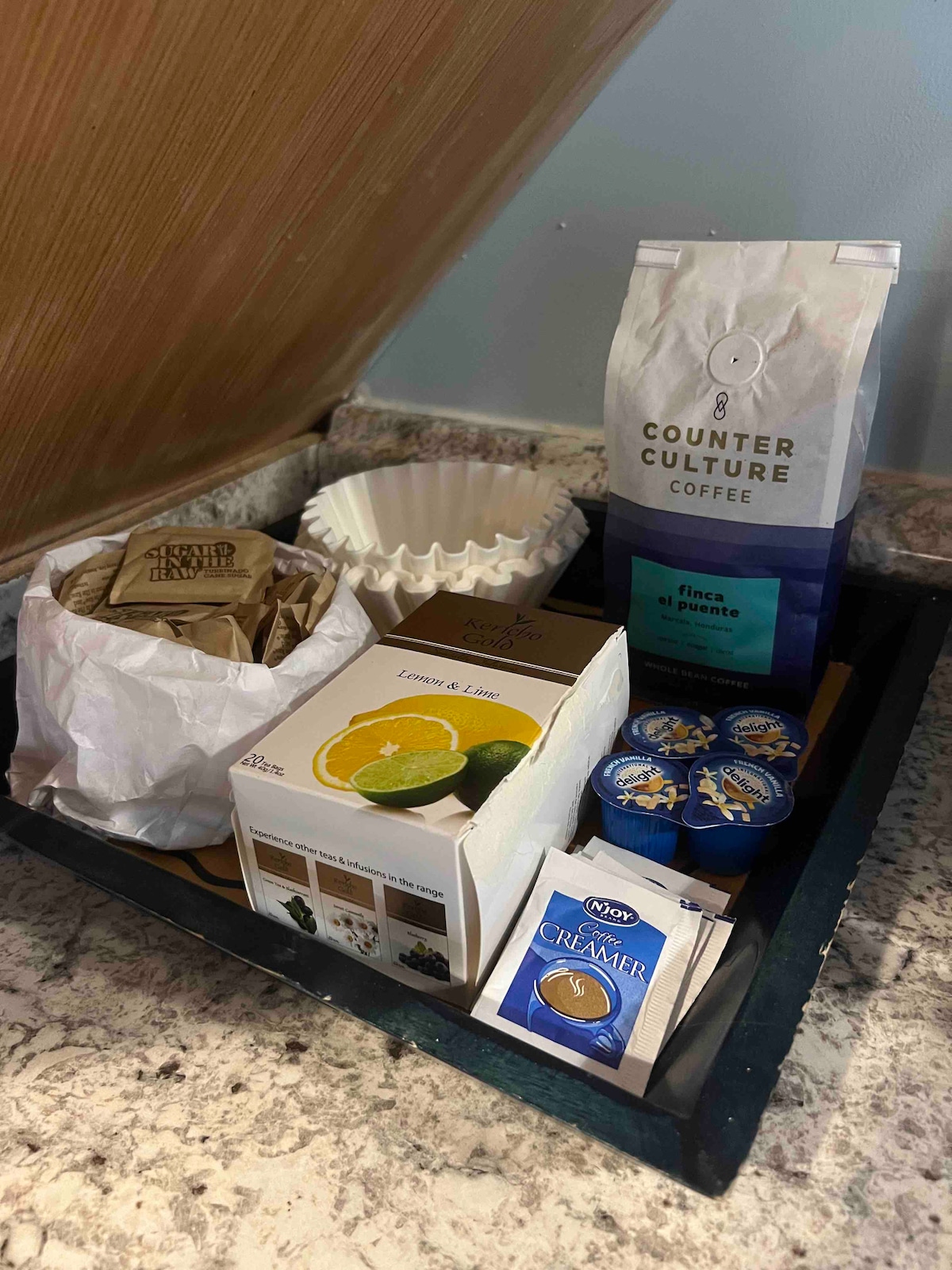 A neatly arranged tray features a selection of coffee supplies, including a bag of Counter Culture coffee, coffee filters, and flavored creamers. An assortment of lemon and lime tea bags is also included, providing a variety of beverage options for guests.