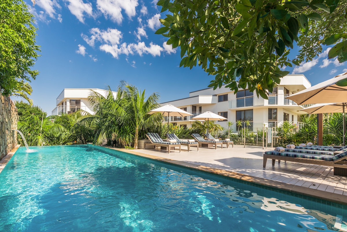 A resort-style pool is surrounded by lush tropical plants and comfortable loungers. Parasols provide shade over the seating areas, and the clear blue water reflects the sunny sky, creating a serene outdoor space for relaxation.