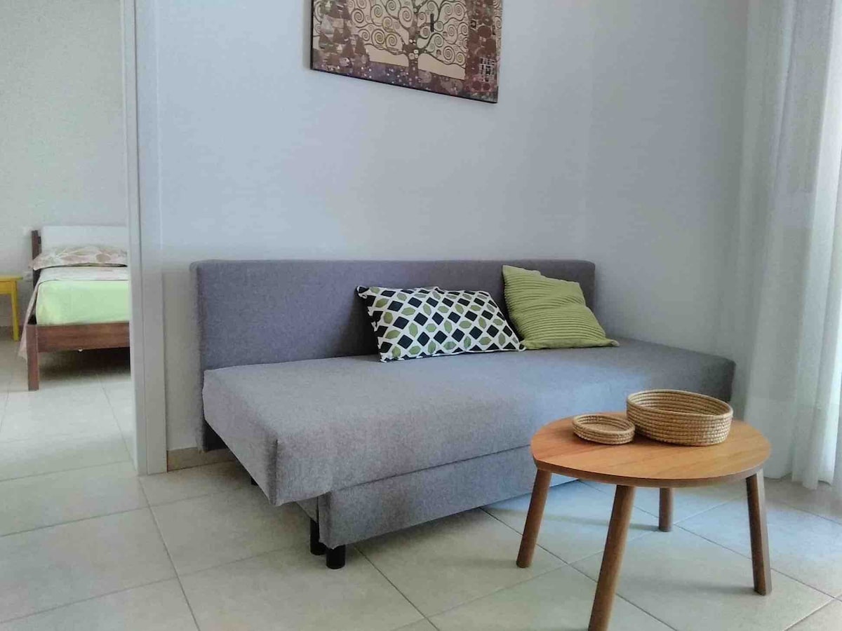 A gray sofa bed is positioned beside a round wooden table with two woven baskets. A subtle artwork is displayed on the wall above, while a softly lit sleeping area is visible in the background. Natural light filters in through sheer curtains.