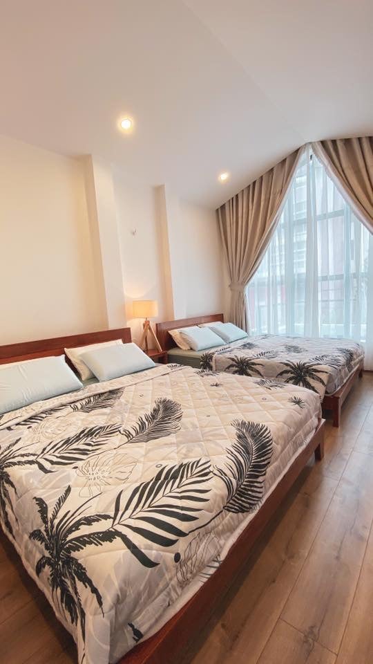 The room features two spacious beds adorned with tropical-themed bedding. Large windows allow natural light to illuminate the space, which is complemented by soft curtains. A bedside lamp adds a touch of functionality, enhancing the overall comfort of the room.