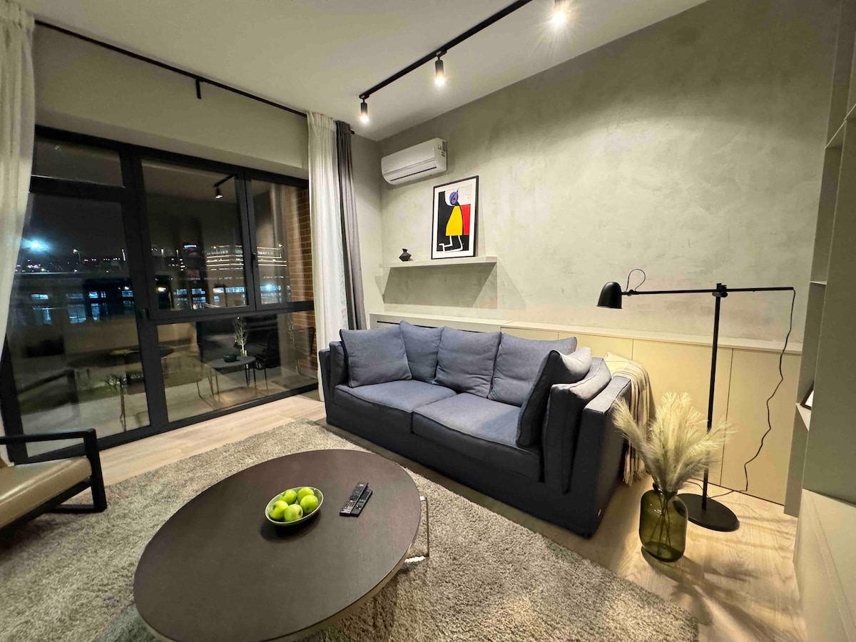 A modern living area is featured with a comfortable blue sofa, a round coffee table with green apples, and a sleek floor lamp. Large glass doors lead to a balcony, allowing natural light to fill the space, while soft curtains frame the view.