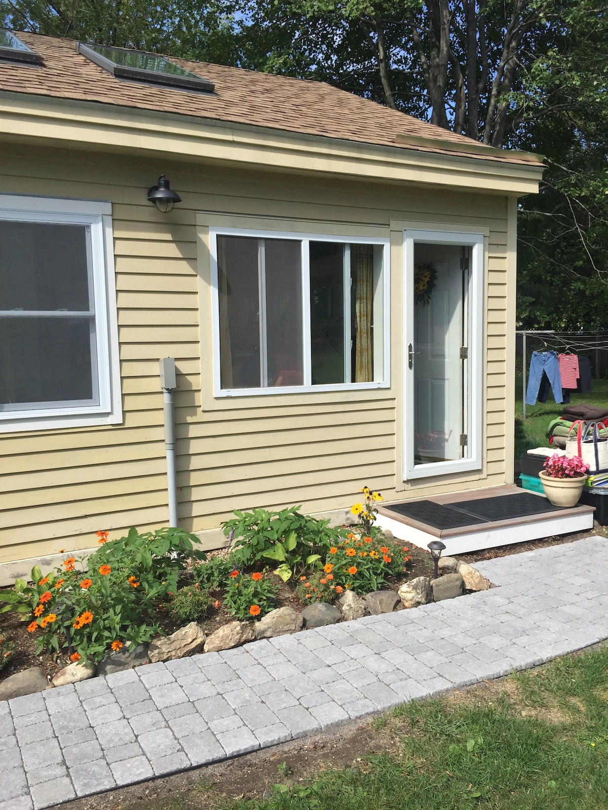 The entrance to a light-colored studio apartment is framed by a stone pathway, leading to a white outer door. Colorful flowers line the garden bed, and laundry hangs on a line nearby, enhancing the garden's cheerful ambiance.