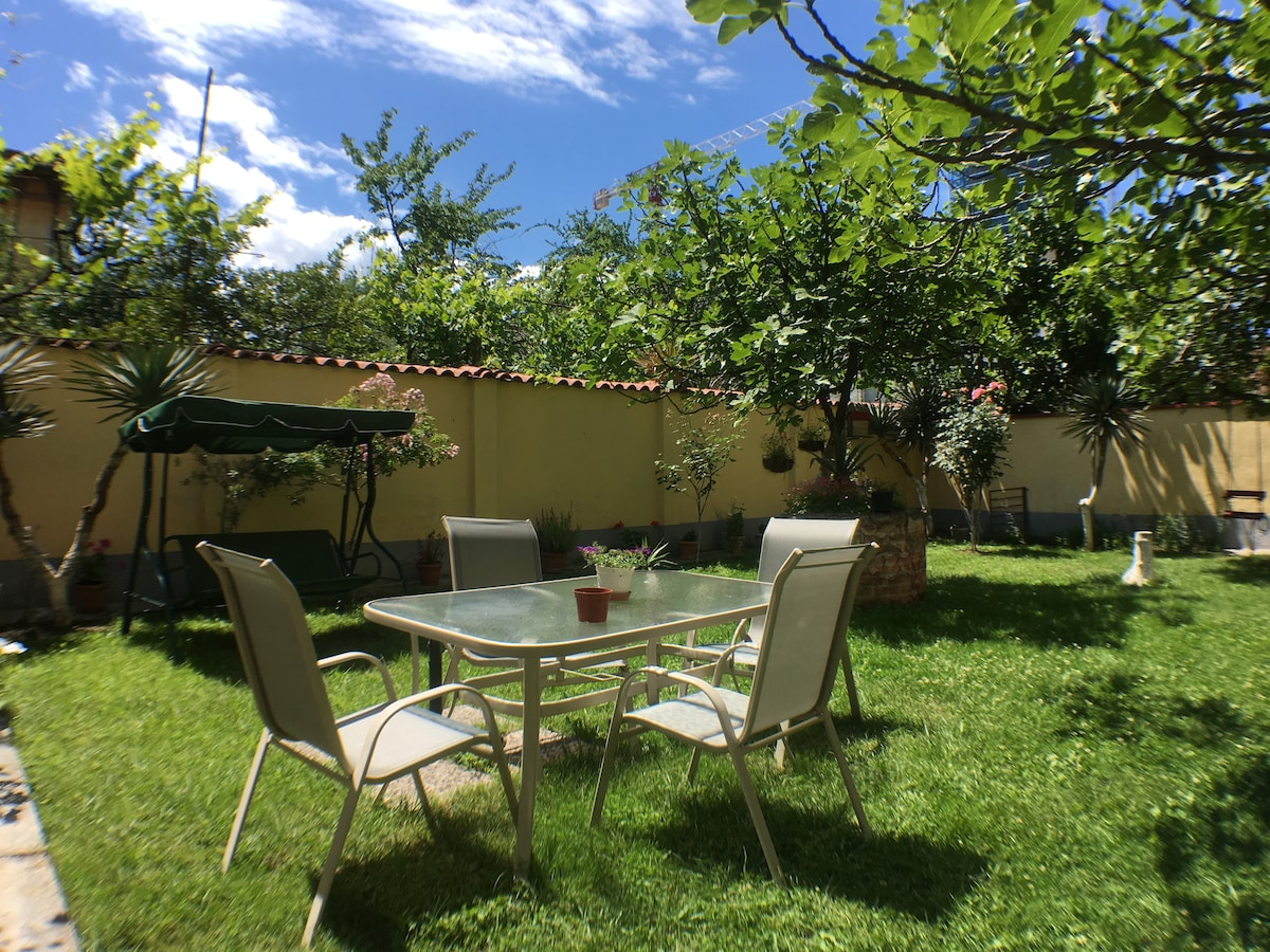 A sunlit garden space features a glass table surrounded by four metal chairs. Lush green grass covers the ground, complemented by various plants and trees in the background. A swing seat adds to the relaxed atmosphere of the private outdoor area.