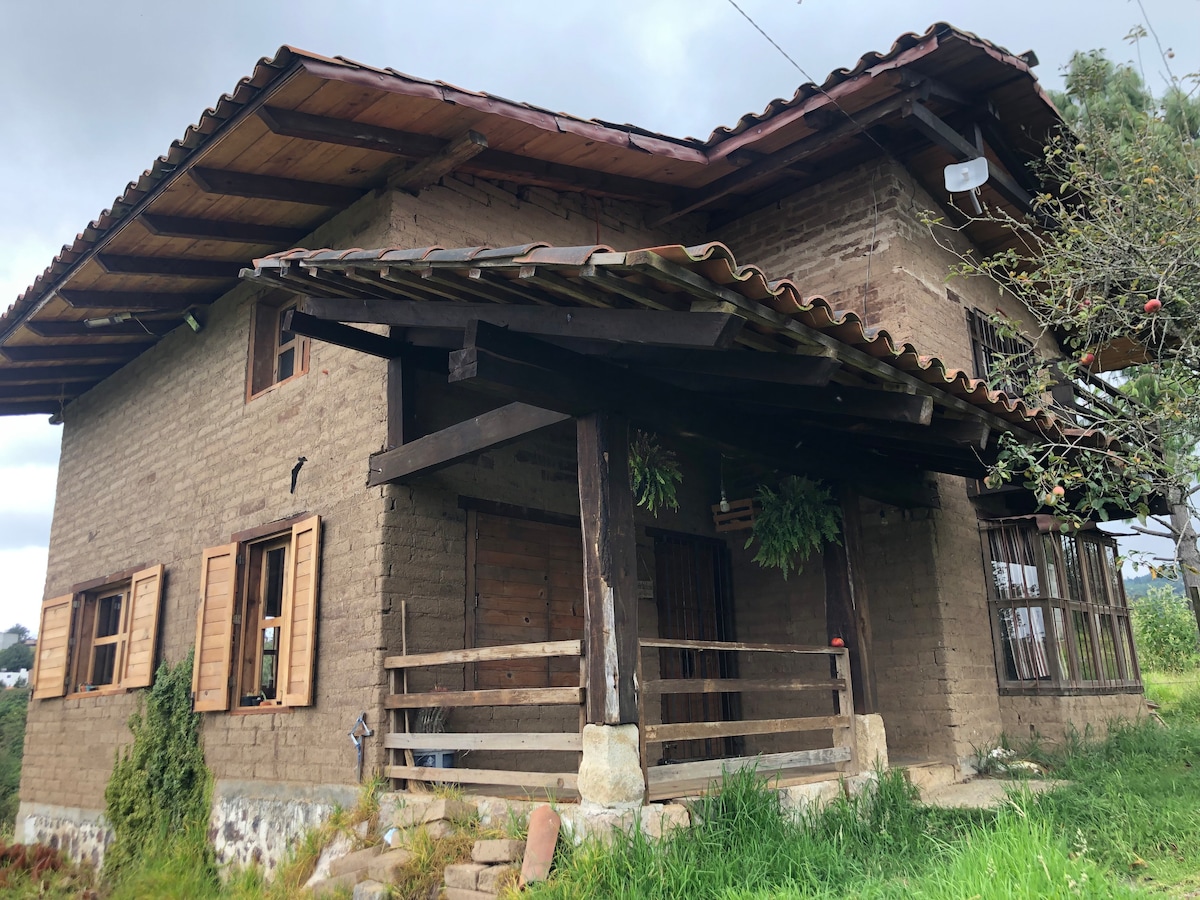 The rustic adobe cabin features a sloped roof with wooden beams and a welcoming porch. Windows with wooden shutters line the facade, allowing ample natural light. Surrounding greenery adds to the tranquil rural setting, enhancing the outdoor space.