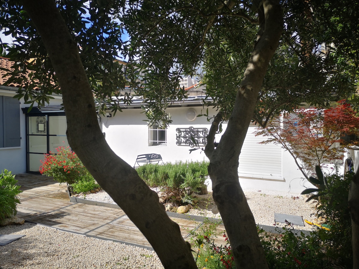 The exterior of a white building is visible, surrounded by greenery and flowering plants. A pathway made of wooden planks leads to the entrance. A small seating area is visible through the foliage, creating a serene and inviting space that connects with nature.