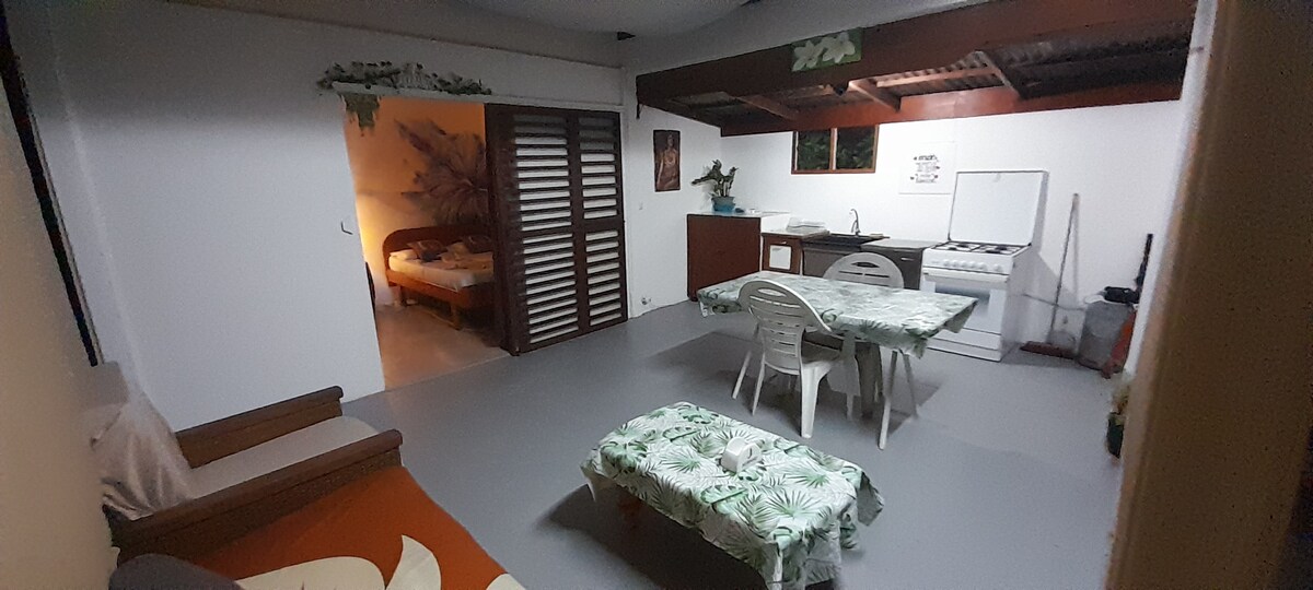 An open living space features a dining area with a table surrounded by chairs. A kitchenette is visible in the background, equipped with cabinets and a stove. A cozy seating area is located to the left, while a bedroom is accessible through sliding doors.