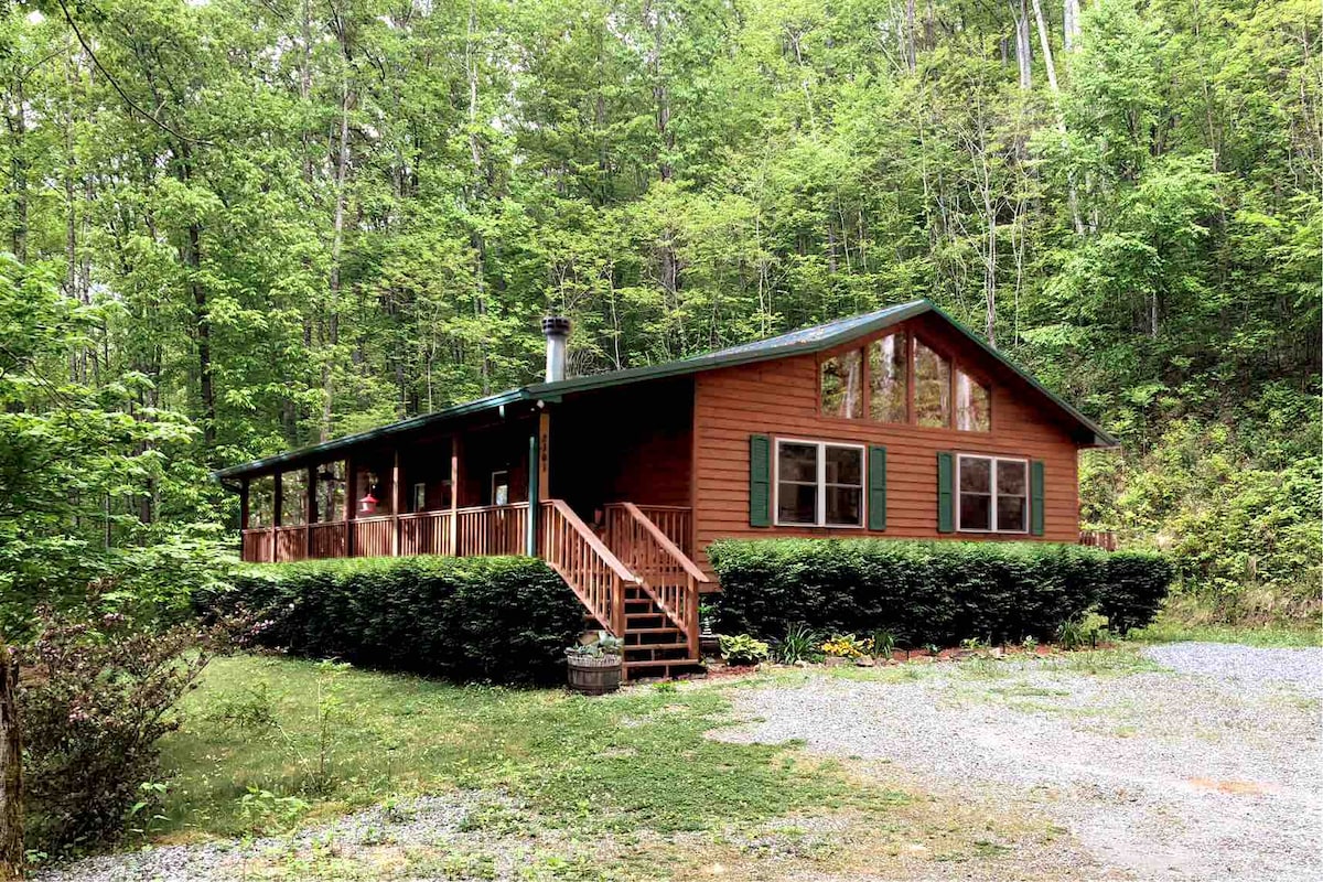 The cabin features a warm wooden exterior surrounded by lush greenery. A spacious front porch with steps leads up to large windows, allowing natural light to fill the interior. A gravel driveway provides access to the entrance, enhancing the cabin's secluded feel.