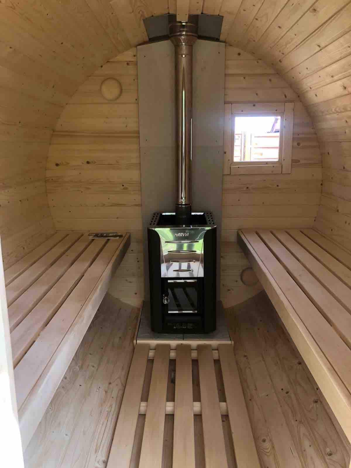 A wooden sauna interior features two wide benches along the walls, crafted from light-colored wood. At the center, a modern wood stove stands on a stone base, complemented by a small window that allows natural light to enter the space.