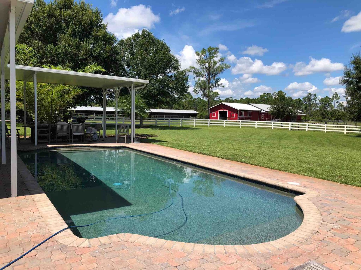 An inviting swimming pool is surrounded by a spacious patio area, featuring a shaded seating arrangement. Lush green grass extends to the background, where a red barn contrasts with the blue sky and fluffy white clouds.
