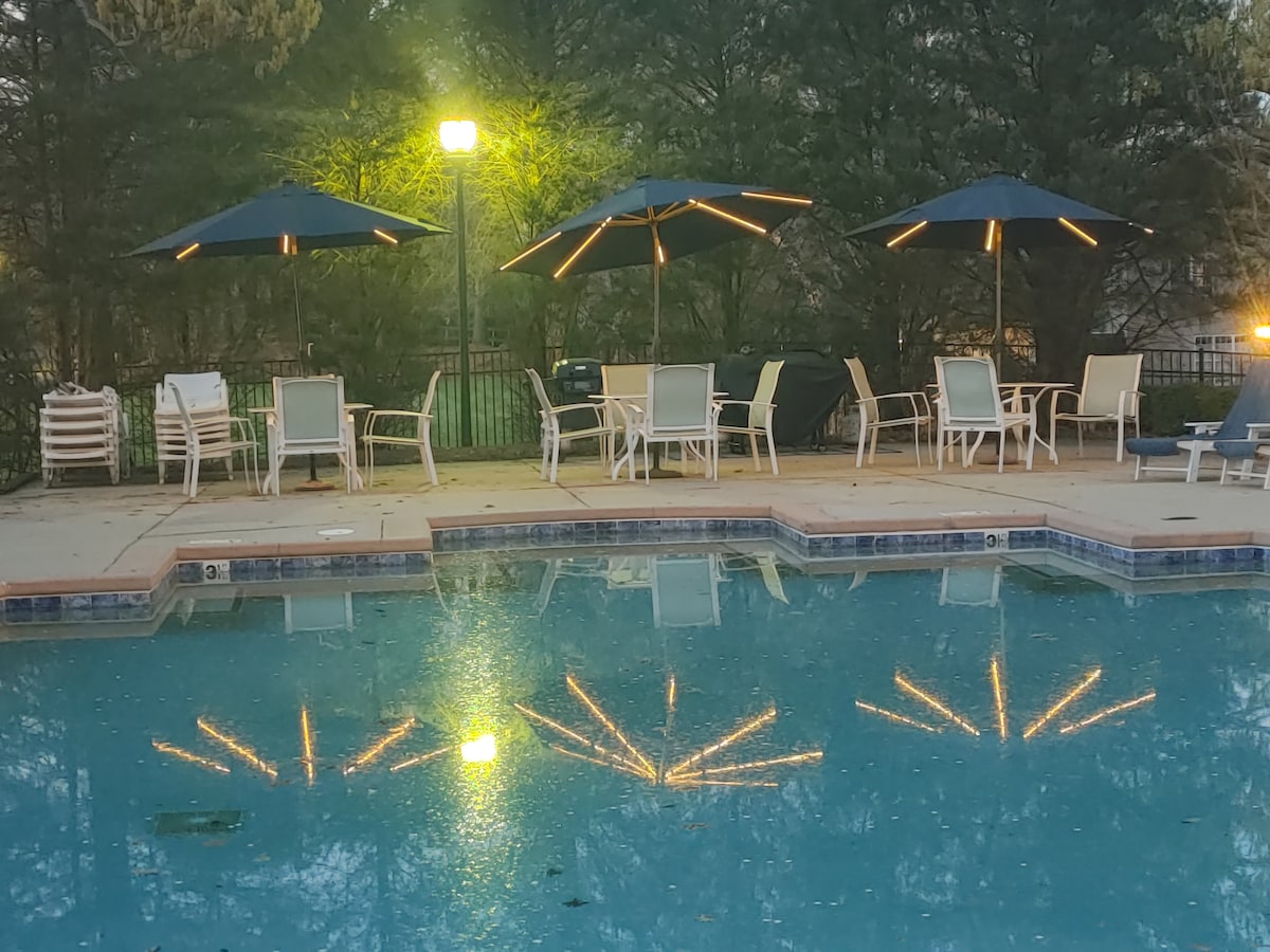 A well-maintained pool area is visible, featuring two large umbrellas providing shade over seating. Lounge chairs are arranged around the pool's edge, with lights reflecting off the water's surface, creating a serene atmosphere. Lush greenery borders the space.