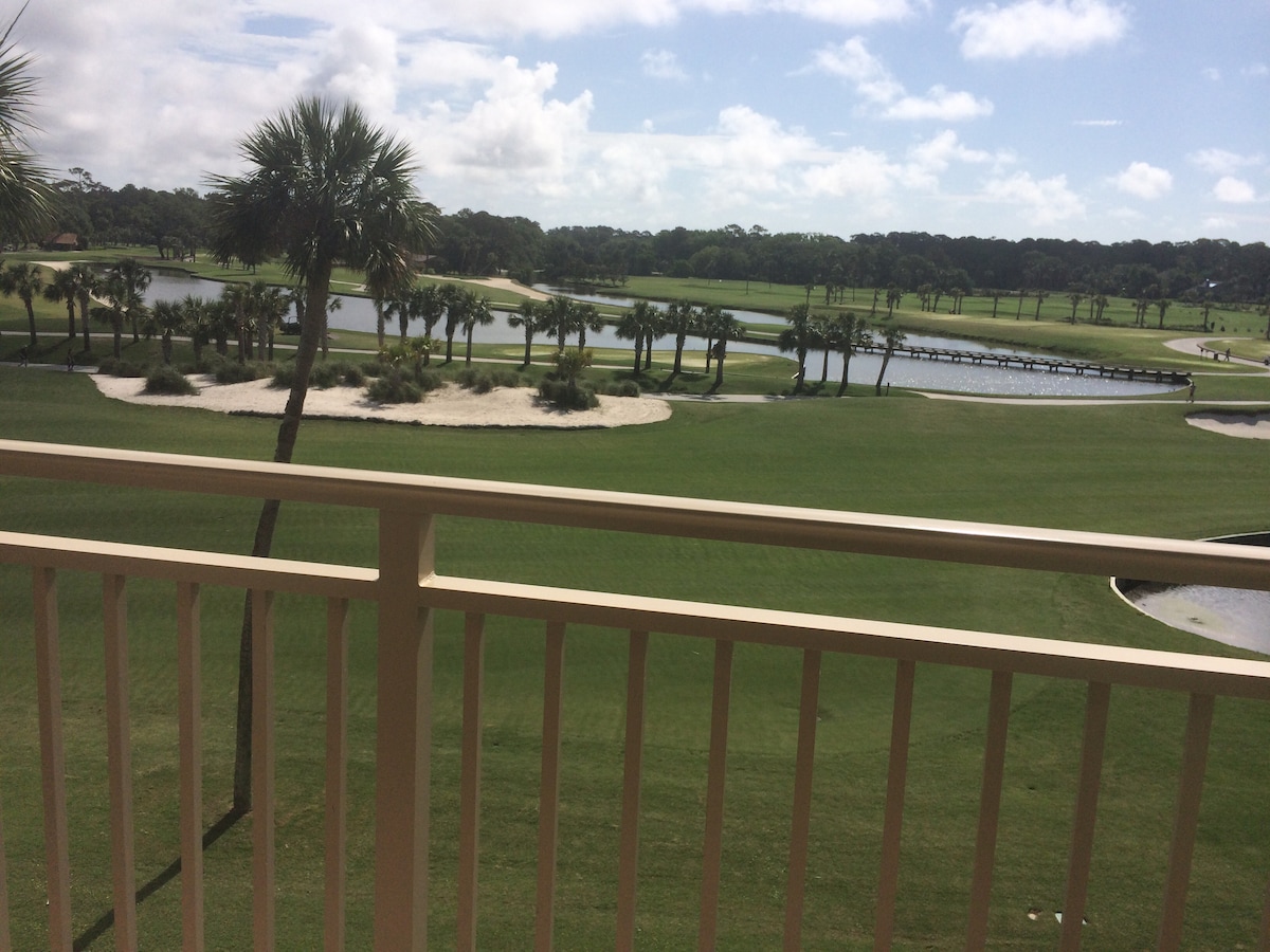 A view of two golf courses and lagoons stretches across the landscape, showcasing well-maintained green spaces and sandy traps. Palm trees are positioned throughout, and blue skies with scattered clouds provide a bright backdrop.