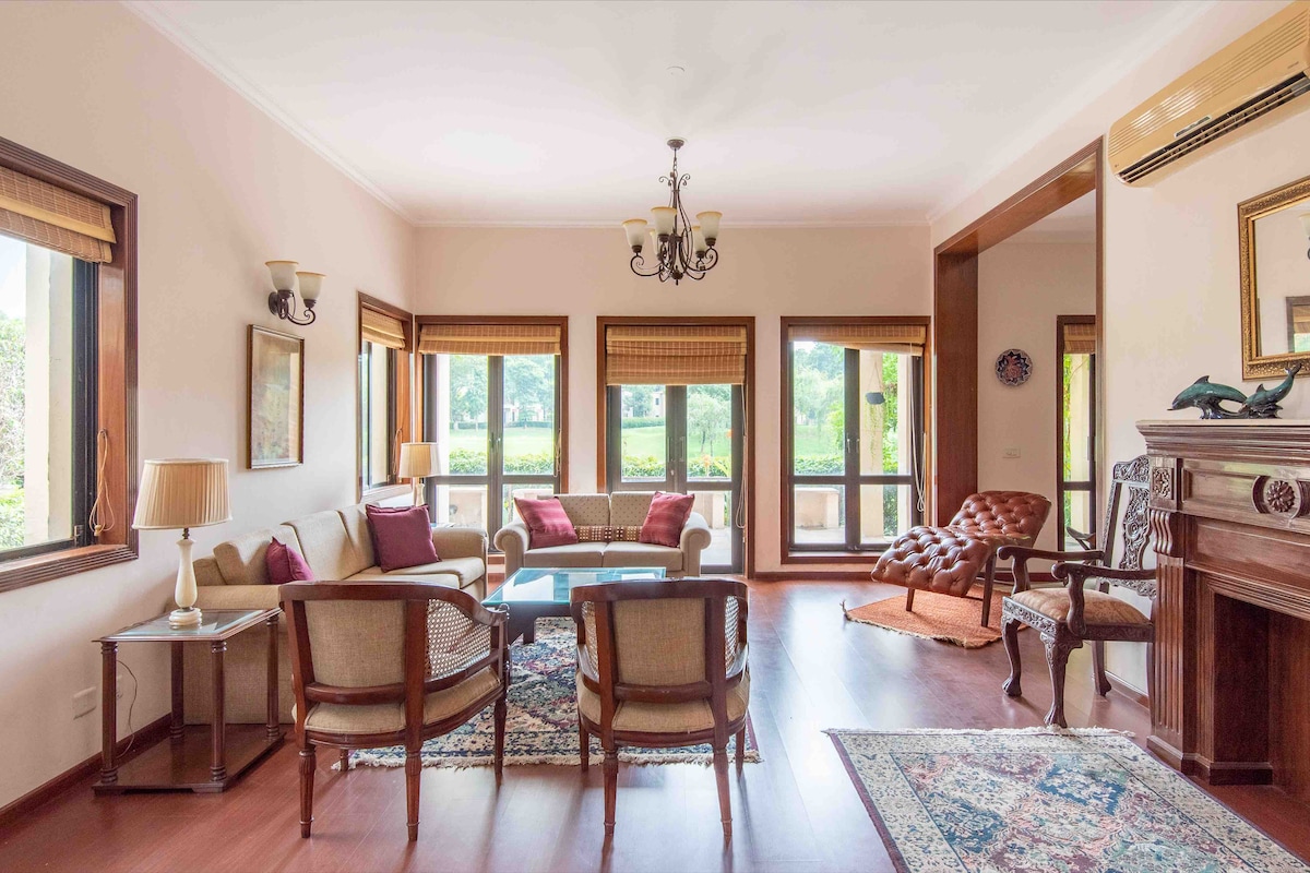 A cozy living area features a blend of modern and traditional decor. Two armchairs and a sofa are arranged around a central coffee table, with warm light filtering through large windows. An elegant chandelier hangs above, and decorative elements add character to the space.