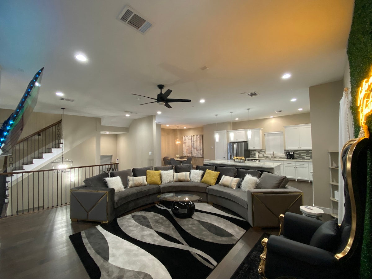 The spacious living area features a circular sectional sofa, accented with decorative cushions, arranged around a central coffee table. The modern kitchen and dining area are visible in the background, with soft lighting enhancing the relaxed ambiance. A dark area rug adds texture to the hardwood flooring.