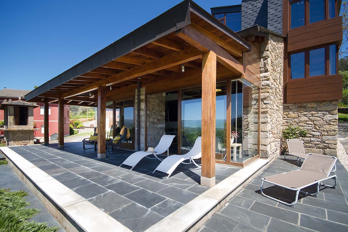 A covered terrace showcases wooden beams and stone accents, providing shade and a relaxed space. Two reclining lounge chairs are positioned on a slate patio, with expansive glass doors offering views of the surrounding landscape. The area is free of clutter, promoting a calm atmosphere.
