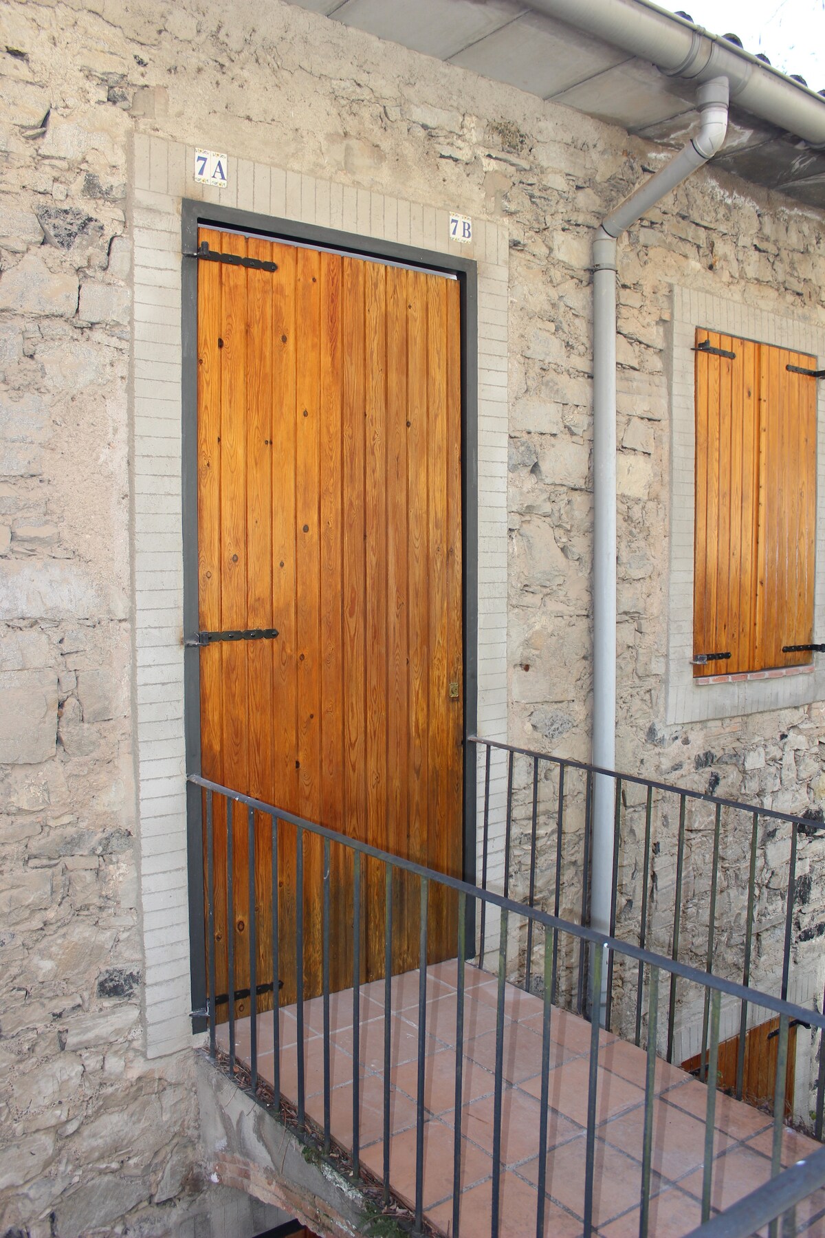 A rustic wooden door features a natural finish, complemented by a black metal frame. The doorway is set against a stone wall, revealing an inviting passage. A small balcony with a black railing leads to the entrance, showcasing the structure's harmonious blend with its natural surroundings.