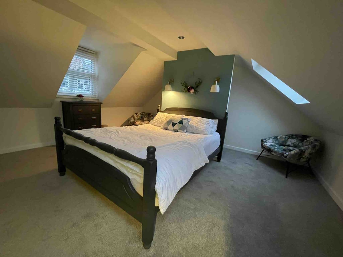 An inviting bedroom features a large bed with a white coverlet, flanked by two bedside lamps. A floral armchair is placed in the corner, and a dresser adds functionality. Natural light enters through a window set in the ceiling's slope.