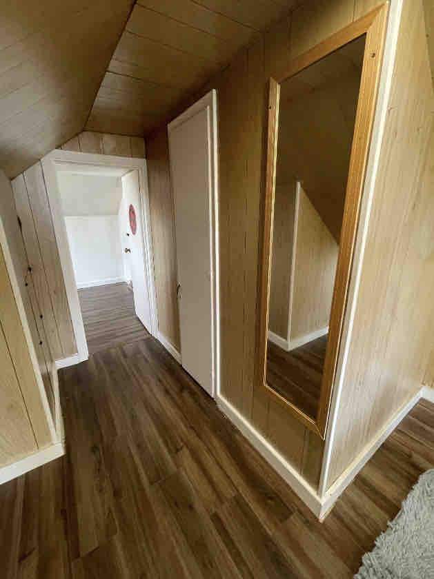 A narrow hallway features warm wooden paneling and laminate flooring. A full-length mirror is mounted on the wall, reflecting light from the nearby space. Doorways lead to other rooms, enhancing the sense of accessibility and openness in the home.