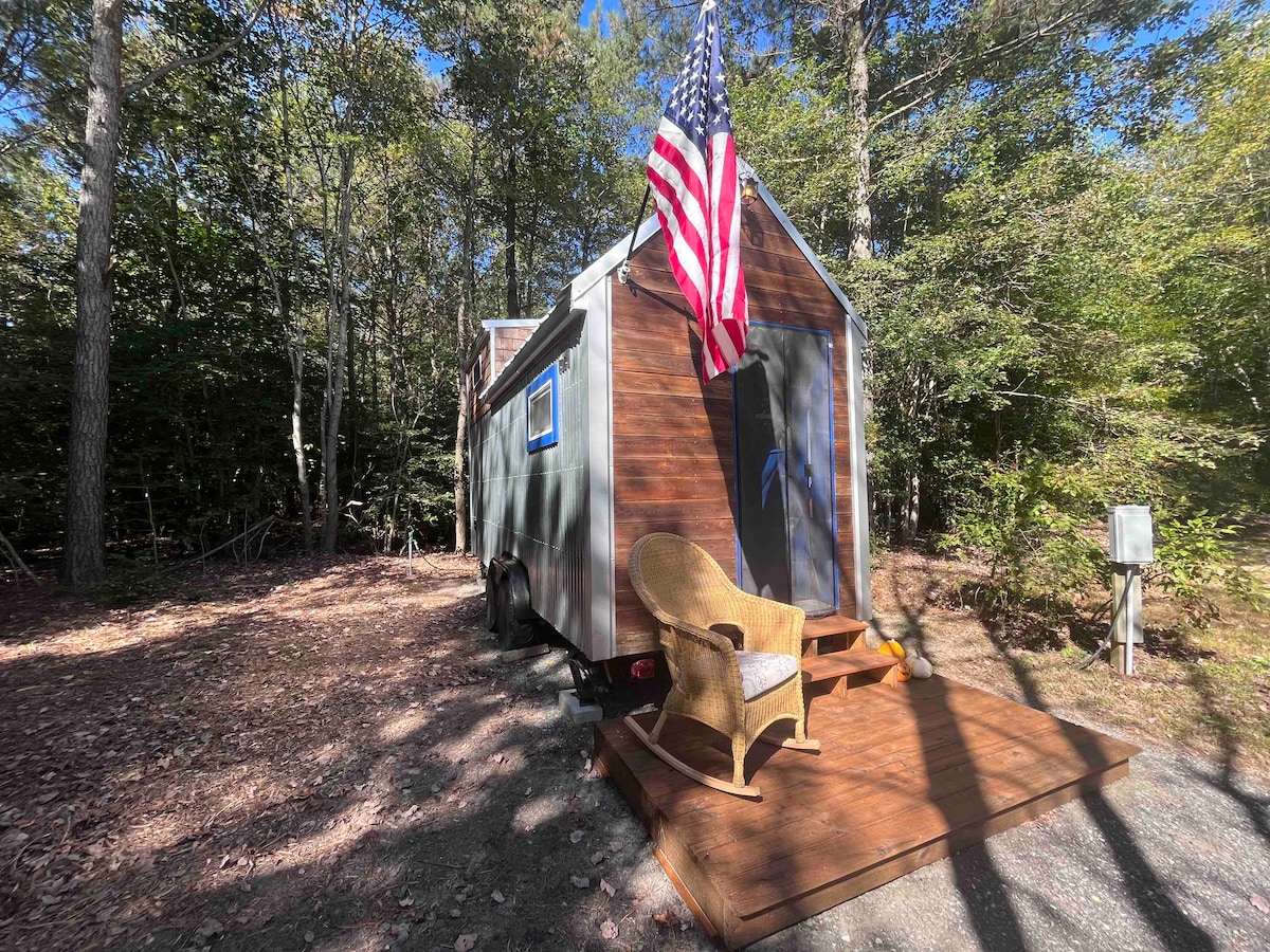 The tiny house is set among a wooded area, featuring a wood deck with an outdoor chair. A flag is displayed prominently at the entrance, with large windows allowing for natural light and views of the surrounding trees.