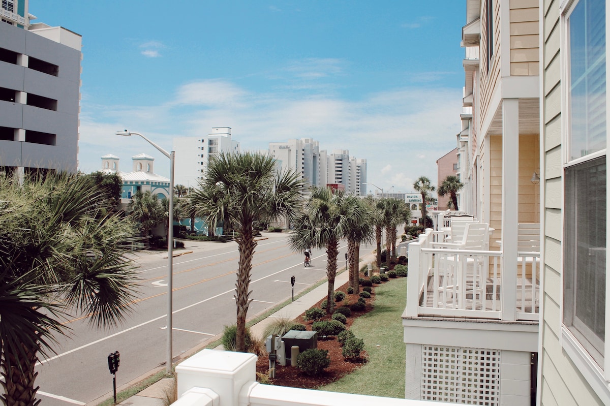 A view from the balcony captures a landscaped area featuring palm trees lining the street. The road leads toward a beachfront area, with low-rise buildings visible in the distance under a clear blue sky.