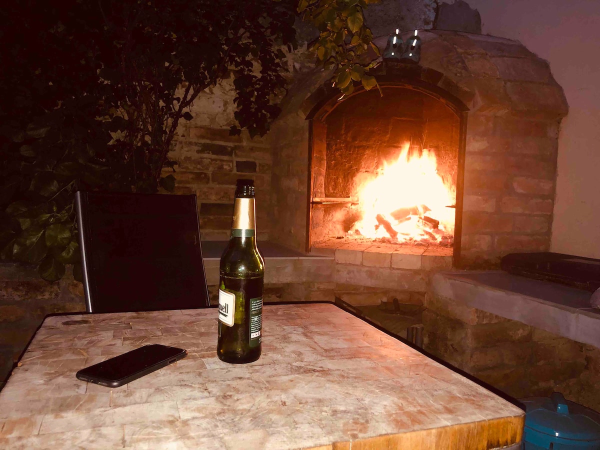 An outdoor area features a stone fireplace, providing a warm glow from a crackling fire. A small table holds a bottle of beer and a smartphone, while lush green plants frame the setting, creating an inviting space for relaxation.