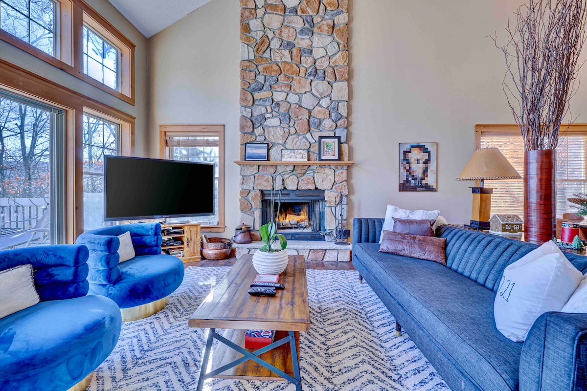 A bright and airy living area features a natural stone fireplace as the focal point. Comfortable blue and gray seating is arranged around a wooden coffee table. Large windows allow natural light to fill the space, complemented by a decorative area rug.