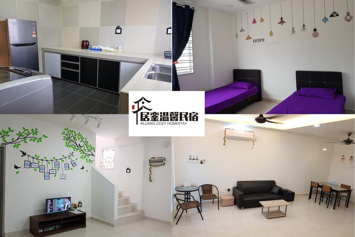 A modern kitchen is equipped with stainless steel appliances and sleek cabinets. Two neatly made beds are dressed in purple linens. A cheerful living area features a comfortable sofa, wall decor with plants, and a television, providing a welcoming ambiance for relaxation.