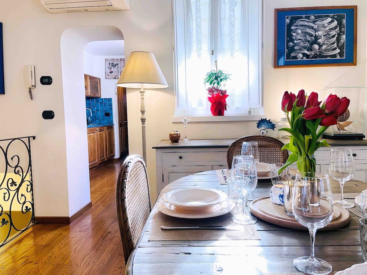 A dining area is presented with a round wooden table set for a meal, featuring plates and glassware. Fresh tulips are arranged in a vase at the center. Natural light filters in through large windows, illuminating the space, with a glimpse of a kitchen visible in the background.