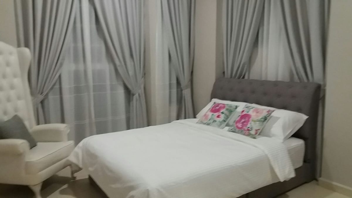 A well-appointed sleeping area features a spacious bed with floral accent pillows, complemented by softly draped curtains. A cozy armchair sits nearby, adding to the comfort of the space. Natural light filters through the windows, enhancing the serene atmosphere.