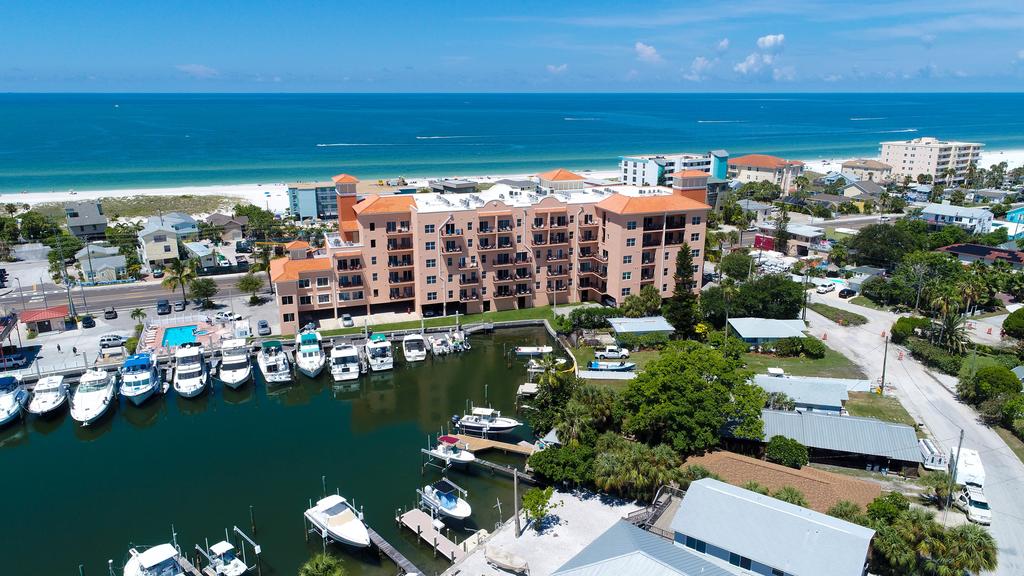 An aerial view captures the Madeira Bay Resort, featuring a six-story building with orange-roofed balconies. The ocean is seen in the distance, alongside a marina filled with boats. Lush greenery surrounds the area, creating a serene atmosphere near the beach.