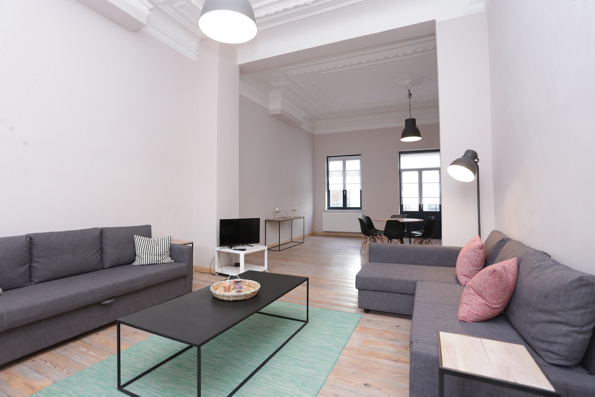 A spacious living area is furnished with two gray sofa sets, complemented by a black coffee table. Large windows allow natural light to fill the room, highlighting the light-colored walls and wooden flooring. A dining area is visible in the background, anchored by a simple table and chairs.