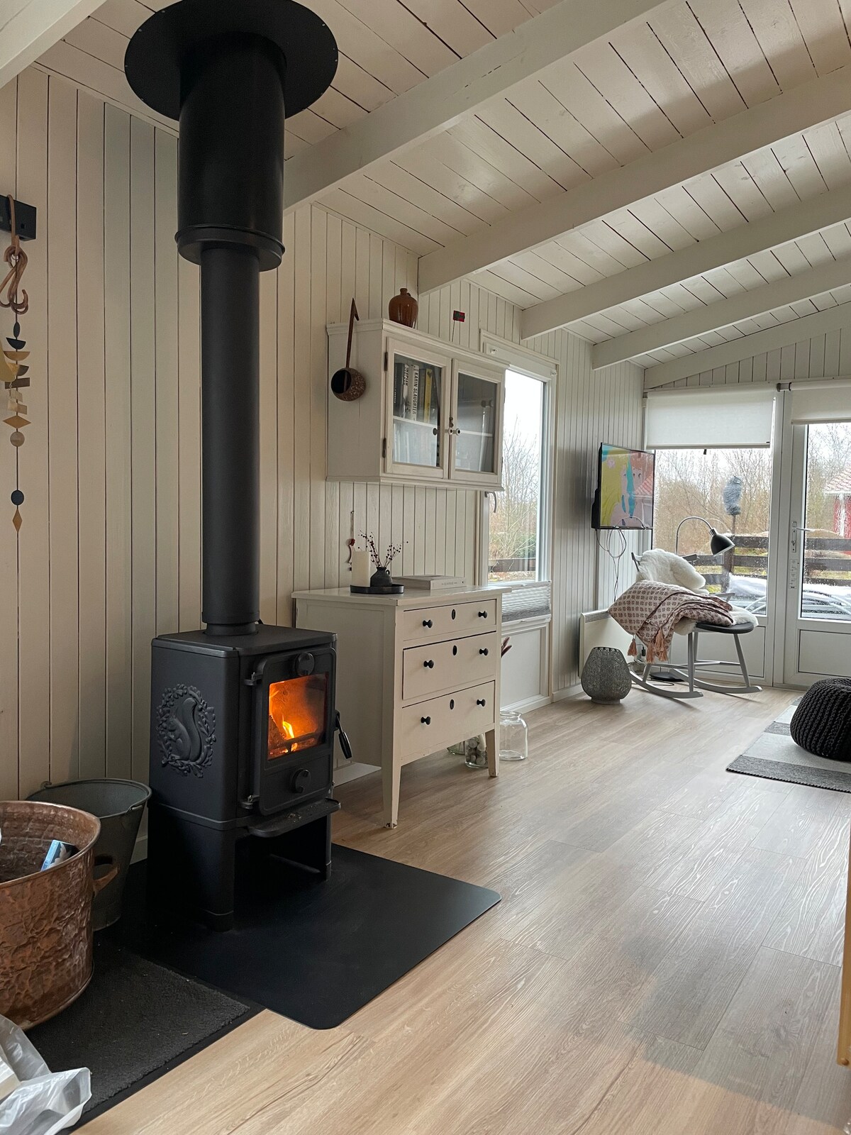 A cozy living space features a modern wood-burning stove, providing warmth and a focal point. Light wood paneling and a white dresser create a bright atmosphere. A spacious armchair and large windows allow natural light to fill the room, with glimpses of the outdoors visible.