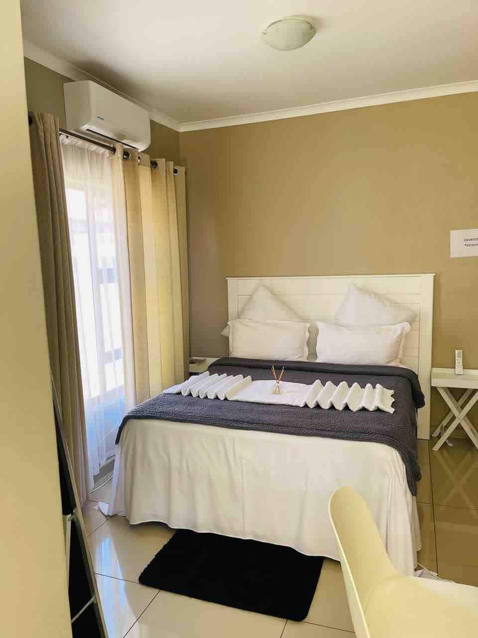 The room features a spacious bed dressed in crisp white linens, accompanied by two pillows. A gray throw blanket is positioned at the foot of the bed. Soft natural light enters through sheer curtains, complementing the neutral wall color. A small table and chair are also visible.