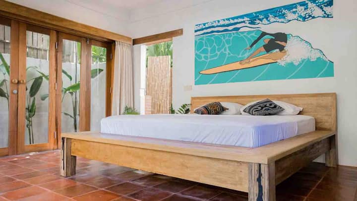 Grand floor bedroom featuring a garden view. The wall features a hand painted mural by a local artist. The room is fitted with AC, seating and desk, a comfy bed and clothes storage.