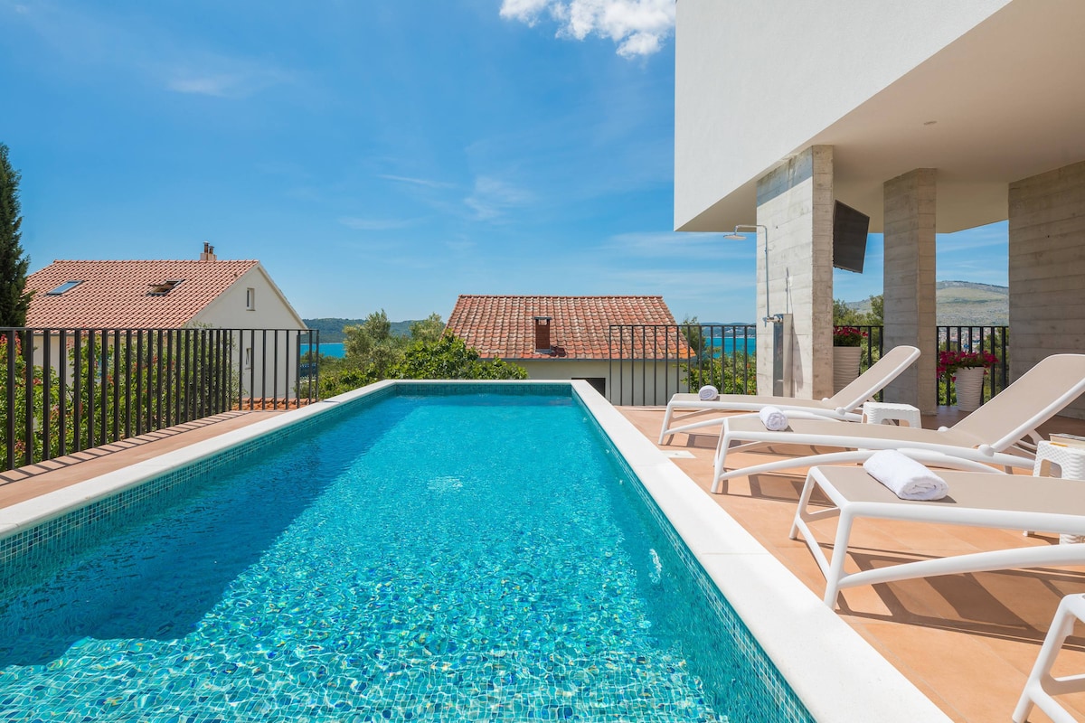 A modern swimming pool is framed by a spacious deck area, featuring white lounge chairs with rolled towels. The pool reflects the clear blue sky, with views of nearby rooftops and greenery visible in the background.