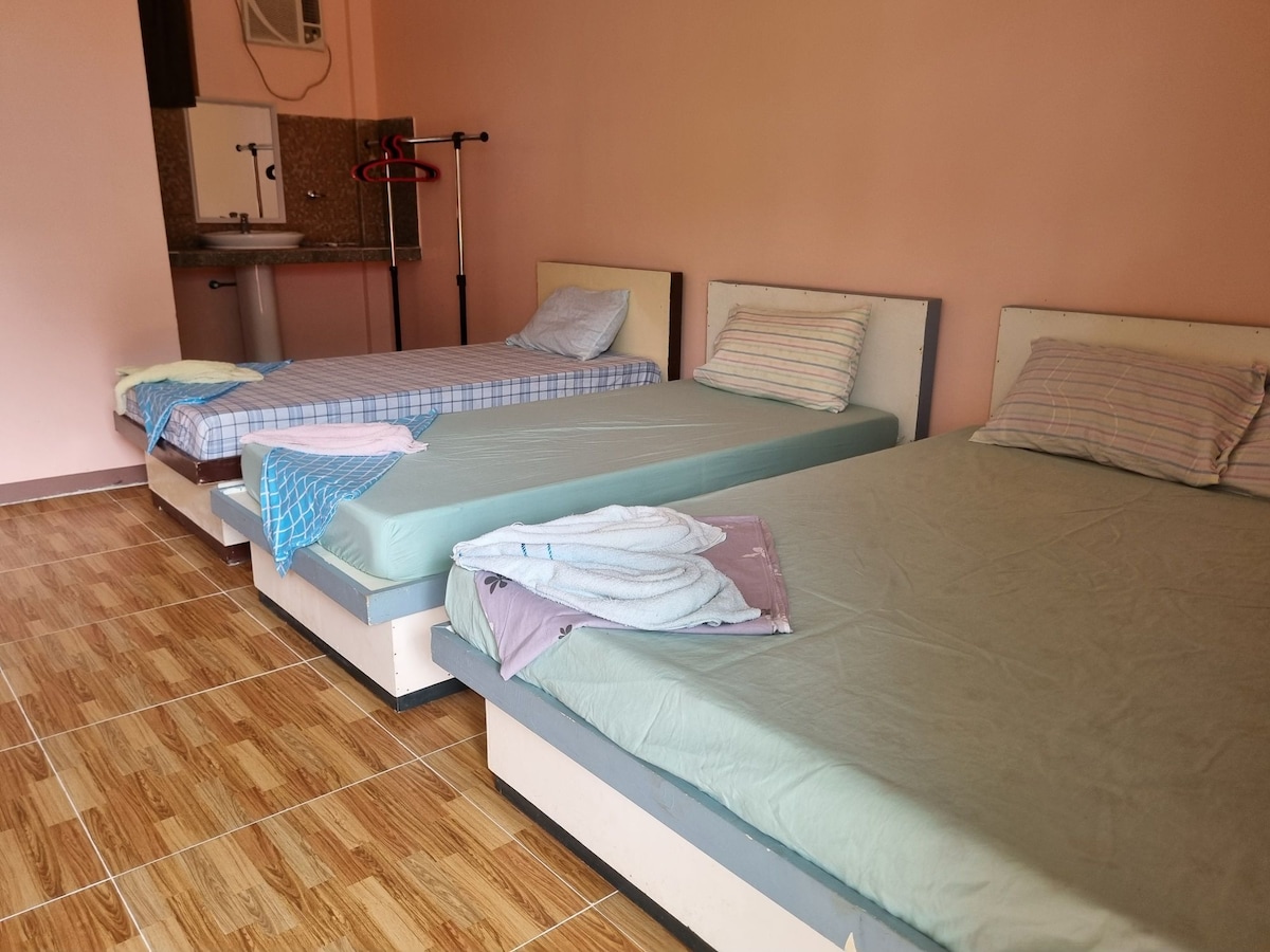 Three spacious beds are arranged neatly in the bright room, each covered with a fitted sheet and a colorful blanket. A kitchenette is visible in the background, along with light-colored walls and tiled flooring, creating a clean and open environment.