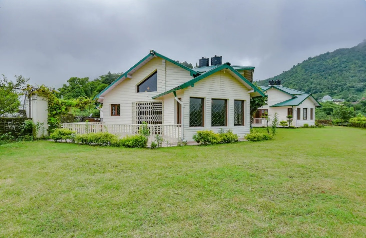 A vintage property is set on a spacious lawn, featuring two villas with sloped roofs and a mix of large windows. Surrounding greenery contributes to a serene atmosphere, while the misty mountains are visible in the background. A harmonious blend with nature is evident.