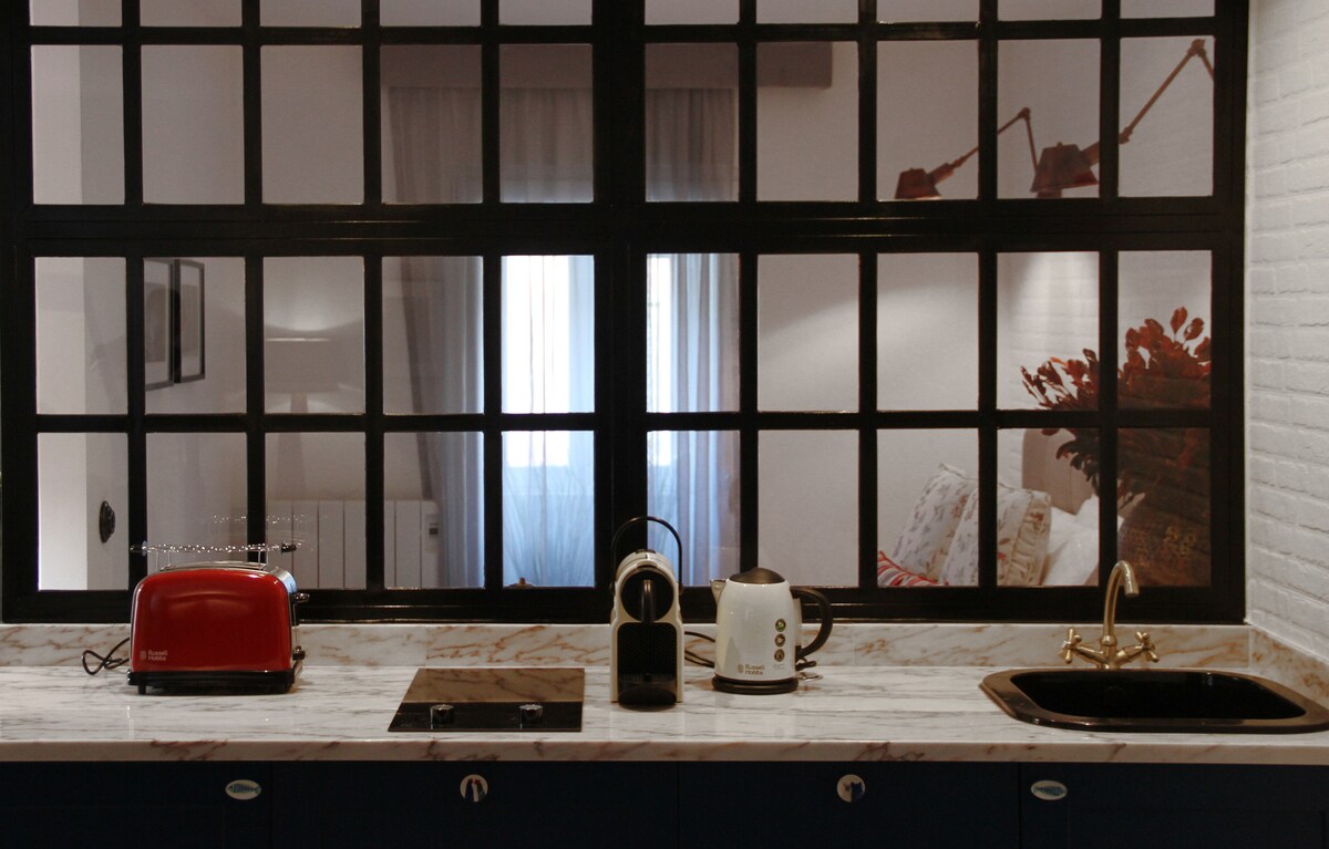 A modern kitchen area features a marble countertop with a red toaster, coffee maker, and kettle. A dark sink is positioned next to a built-in stovetop. Large windows allow light to fill the space, revealing soft furnishings in the adjacent living area.