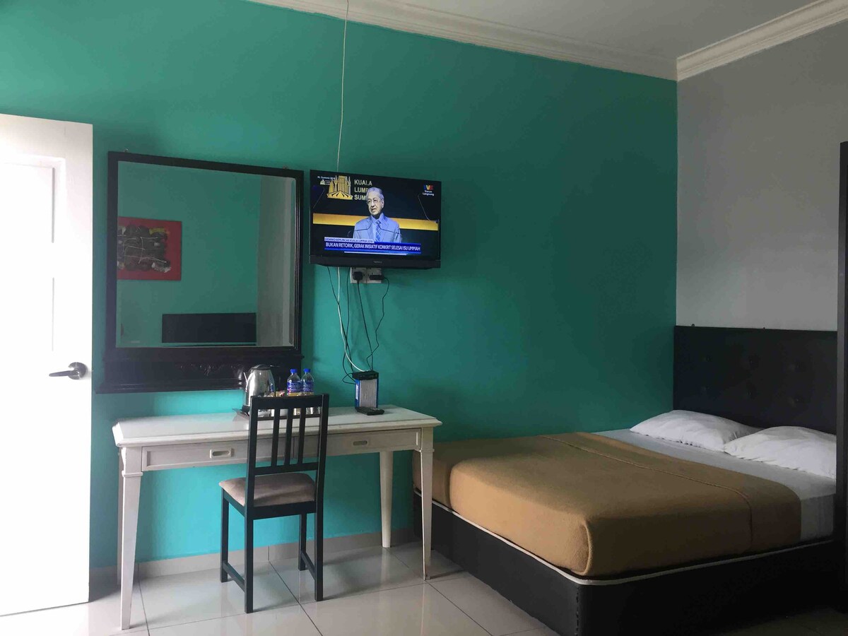 A room with a turquoise accent wall features a spacious bed with neutral bedding. A desk and chair are positioned in front of a mirror. A flat-screen TV is mounted on the wall, while a small refrigerator is visible on the counter.