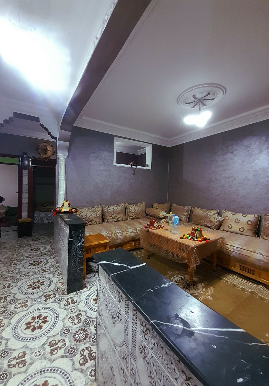 A cozy living area features a large L-shaped sofa adorned with decorative cushions. A wooden table sits in the center, accented with floral arrangements. The space is framed by intricately designed walls and a traditional ceiling, complemented by patterned flooring.