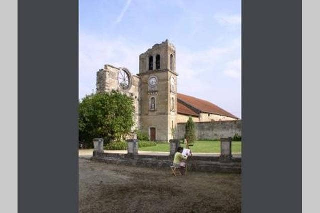 The remains of a historic church building are depicted, featuring a partially collapsed clock tower. The structure is set against a clear sky, with greenery surrounding the area. A figure is seen sitting on a chair, engaging with the surroundings.