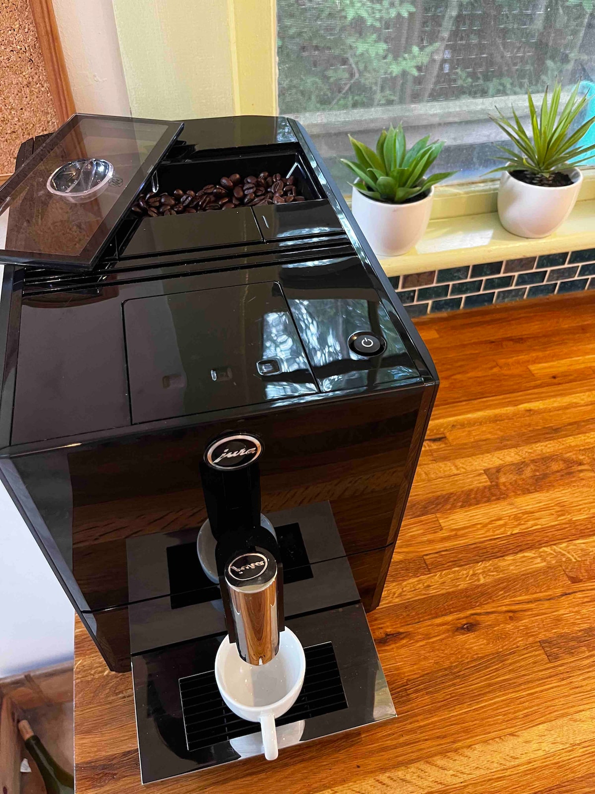 An espresso machine is positioned on a wooden counter, featuring a black exterior and a transparent lid revealing coffee beans. Two coffee dispensers are visible below, with white mugs placed to catch the brewed coffee. Natural light filters through a window, highlighting surrounding plants.