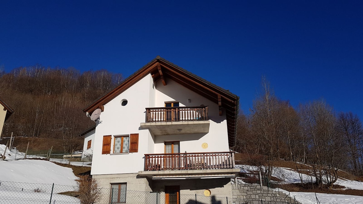 The exterior of the building is showcased, featuring a two-story structure with wooden balconies and large windows. Snow-covered ground surrounds the house, with a backdrop of bare trees and a clear blue sky. The architecture reflects a mountain retreat aesthetic.