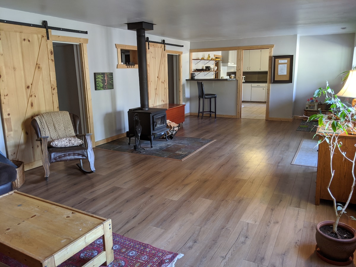 An open living space features a wooden floor and natural light streaming in. A cozy seating area includes a chair and a small table by a decorative rug. A wood-burning stove is centrally positioned, while the kitchen area is visible in the background.