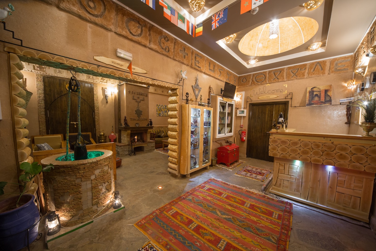 The reception area features a decorative stone fountain at its center, surrounded by intricate murals and artifacts. Wooden accents and a colorful rug enhance the warm ambiance, while flags from various countries are displayed overhead, welcoming guests into the space.