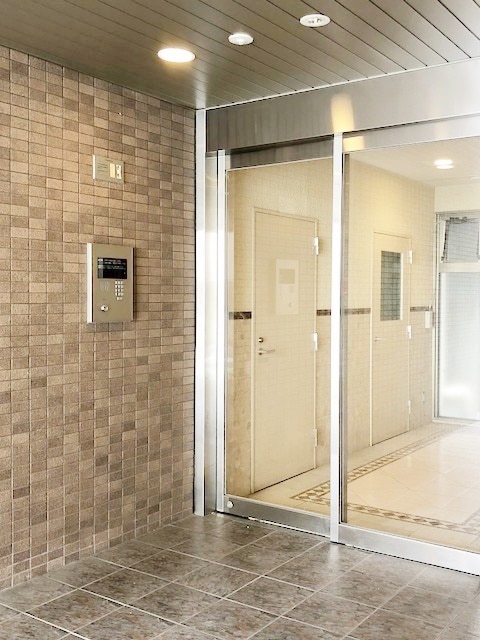 The entrance features a sleek glass door alongside an intercom system. Neutral-toned tile walls and flooring create a modern look, while soft lighting enhances the welcoming feel of the entryway.